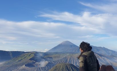 person in jacket sitting on railing with mount bromo and volcanoes behind