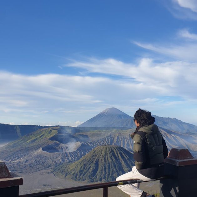 person in jacket sitting on railing with mount bromo and volcanoes behind