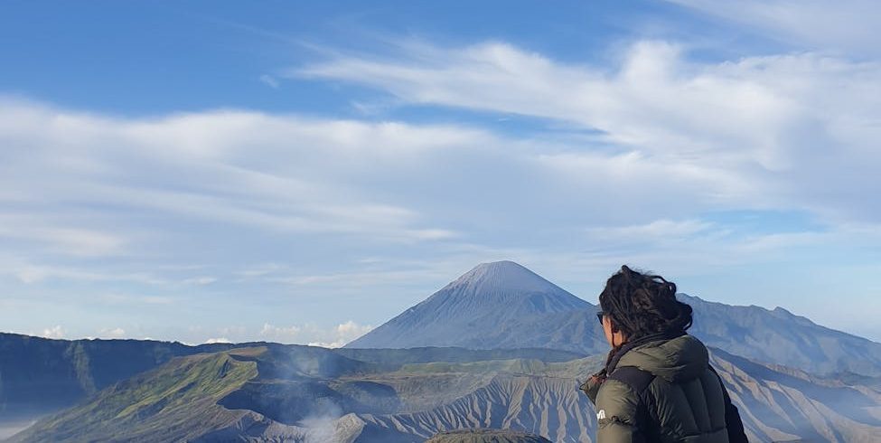 person in jacket sitting on railing with mount bromo and volcanoes behind