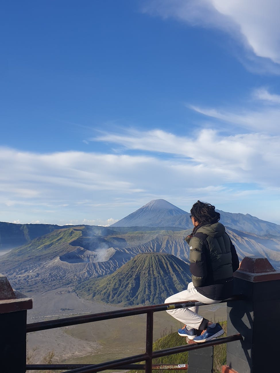person in jacket sitting on railing with mount bromo and volcanoes behind